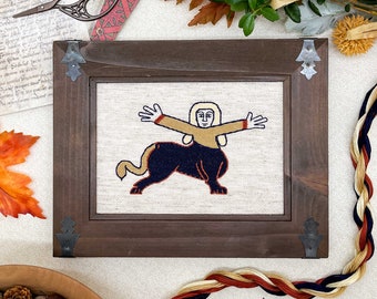 May include: A framed cross-stitch embroidery depicting a mythical creature with a human torso and a lion's body. The creature is depicted in a beige, brown, and blue color scheme. The embroidery is framed in a dark brown wood frame with metal accents.
