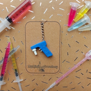 May include: A blue miniature pipette keyring with the word "POSH" printed on it, hanging on a brown tag. The background includes syringes and test tubes filled with colourful liquids, and the text "myMini_aboratory".