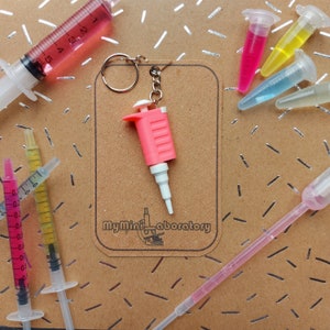 May include: A pink and white miniature pipette keychain with a silver keyring. The keychain is surrounded by syringes and small vials filled with colorful liquids. The text "MyMiniLaboratory" is visible on a brown background.