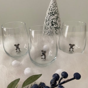Può includere: Tre bicchieri da vino in vetro trasparente con una piccola statuina di koala grigia all'interno di ciascuno. I bicchieri sono appoggiati su una superficie bianca e soffice con un piccolo albero di Natale artificiale bianco sullo sfondo.