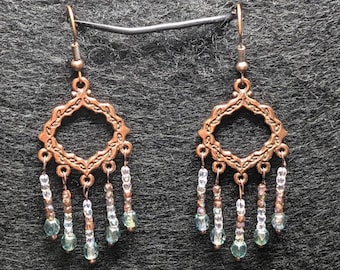 May include: A pair of copper-colored earrings with a diamond-shaped filigree design. Each earring has dangling strands of small, iridescent blue and clear beads, adding a touch of sparkle. The earrings are suspended from a simple hook.