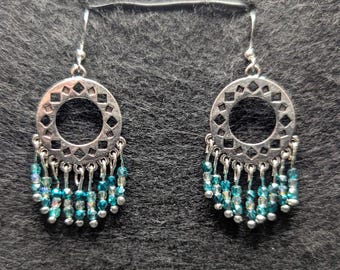 May include: A pair of silver-toned dangle earrings. Each earring features a circular design with geometric cutouts and a fringe of teal and silver beads. The earrings hang from a simple hook.