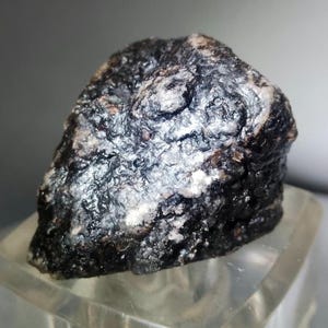 May include: A dark gray, metallic meteorite with a rough, textured surface. The meteorite has a silvery sheen and appears to be covered in small, white crystals.