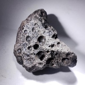 Peut inclure: Une roche sombre et poreuse, parsemée de nombreux trous de tailles variées. La surface de la roche est rugueuse et texturée, suggérant un fragment. L'éclairage met en évidence la forme irrégulière et les différentes tailles des trous.