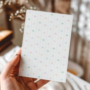 May include: A rectangular card with a repeating pattern of colourful bows and dotted lines on a white background. The bows are in shades of pink, green, yellow, and purple. The card is held by a hand and is likely for stationery or crafts.