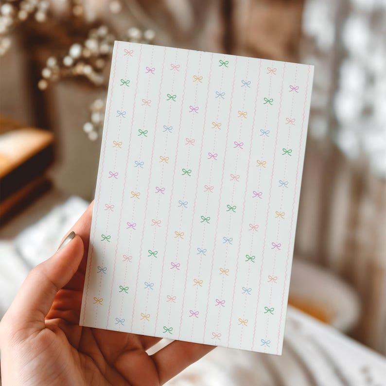 May include: A rectangular card with a repeating pattern of colourful bows and dotted lines on a white background. The bows are in shades of pink, green, yellow, and purple. The card is held by a hand and is likely for stationery or crafts.