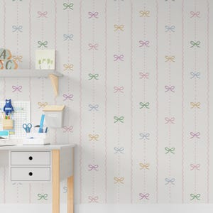 May include: A white wall decorated with a repeating pattern of pastel bows and wavy lines. The bows are in pink, green, yellow, and blue. A desk with a pegboard, shelf, and a small mailbox is in the foreground.