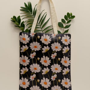 May include: A white canvas tote bag with natural cotton straps. The bag features a black background with a repeating pattern of white daisies with orange centers.