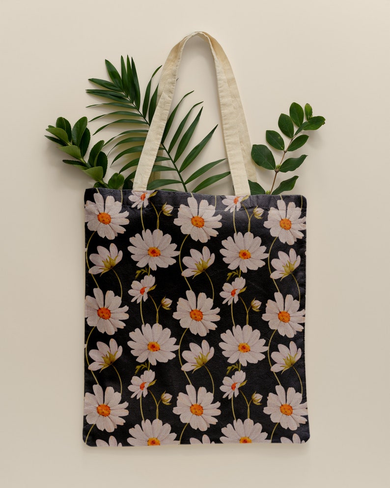 May include: A white canvas tote bag with natural cotton straps. The bag features a black background with a repeating pattern of white daisies with orange centers.