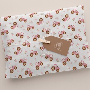 May include: Wrapped gift with a light gray background. The wrapping paper features a repeating pattern of pink tractors and small pink flowers. A brown tag with the words "Happy Birthday" is attached with a wooden clip and twine.