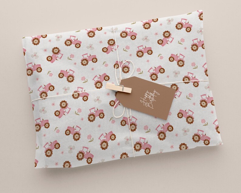 May include: Wrapped gift with a light gray background. The wrapping paper features a repeating pattern of pink tractors and small pink flowers. A brown tag with the words "Happy Birthday" is attached with a wooden clip and twine.