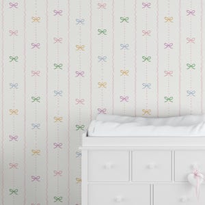 May include: White wallpaper featuring a repeating pattern of multi-coloured bows in pink, yellow, green, and blue. Pink wavy lines and dotted lines connect the bows. A white dresser with a changing table is in the foreground.