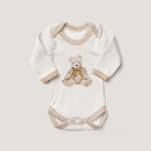 May include: White long-sleeve baby onesie with a brown teddy bear illustration on the front. The onesie has a beige trim around the neckline and snaps at the bottom.