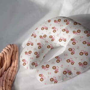 May include: A white nursing pillow with a repeating pattern of pink tractors and brown wheels. The pillow is crescent-shaped and rests on a white surface. A peach-colored, ribbed fabric is visible on the left side of the image.