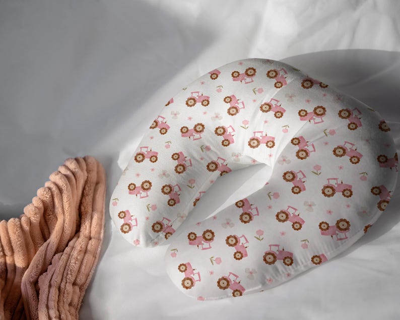 May include: A white nursing pillow with a repeating pattern of pink tractors and brown wheels. The pillow is crescent-shaped and rests on a white surface. A peach-colored, ribbed fabric is visible on the left side of the image.