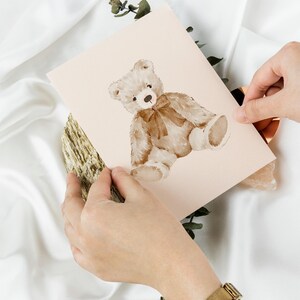 May include: A light pink card with a watercolor illustration of a brown teddy bear wearing a brown bow. The bear is sitting on its hind legs with its paws in front of it.