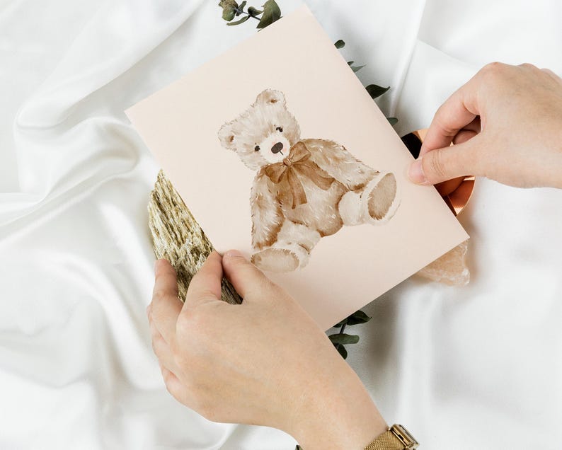 May include: A light pink card with a watercolor illustration of a brown teddy bear wearing a brown bow. The bear is sitting on its hind legs with its paws in front of it.