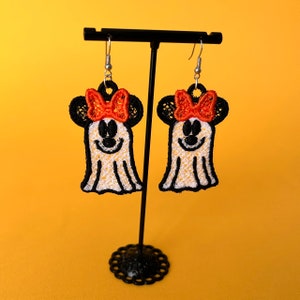Puede incluir: Un par de pendientes con temática de Halloween. Cada pendiente presenta un diseño de fantasma con contornos negros, una cara sonriente y un lazo naranja estilo Minnie Mouse. Los pendientes se exhiben en un soporte negro sobre un fondo amarillo.