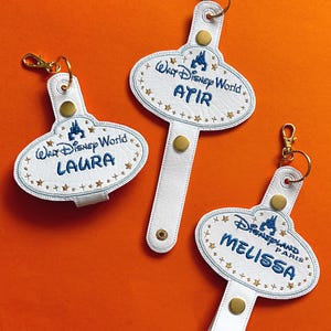 May include: Three white leather keychains with gold hardware and embroidered text. Two say "Walt Disney World" with names "Laura" and "Atir", and one says "Disneyland Paris" with the name "Melissa". All have a blue castle logo and star accents.