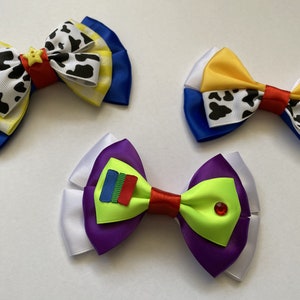 Pu&ograve; includere: Tre fiocchi per capelli fatti a mano a tema Toy Story. Il primo fiocco &egrave; blu, giallo e bianco con una stampa di mucca e una stella gialla. Il secondo fiocco &egrave; blu, bianco e giallo con una stampa di mucca. Il terzo fiocco &egrave; bianco, viola e verde con un rettangolo rosso e blu e un gioiello verde.