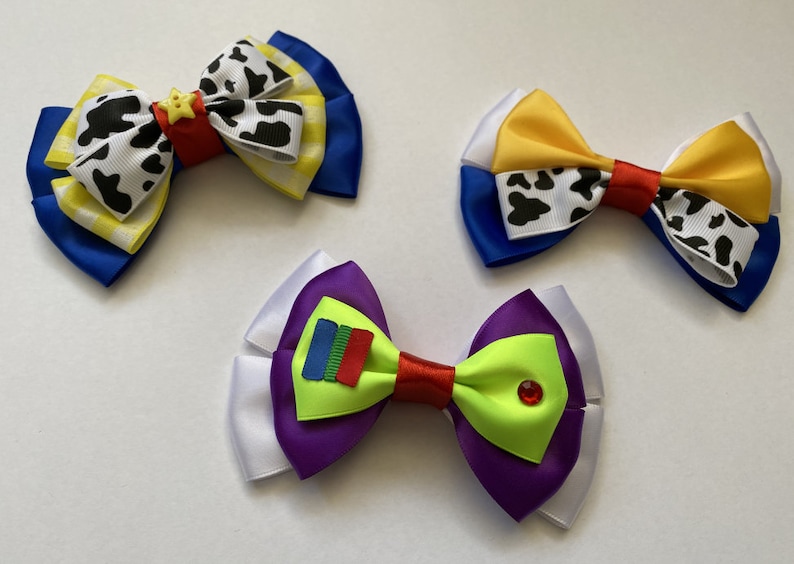 Pu&ograve; includere: Tre fiocchi per capelli fatti a mano a tema Toy Story. Il primo fiocco &egrave; blu, giallo e bianco con una stampa di mucca e una stella gialla. Il secondo fiocco &egrave; blu, bianco e giallo con una stampa di mucca. Il terzo fiocco &egrave; bianco, viola e verde con un rettangolo rosso e blu e un gioiello verde.