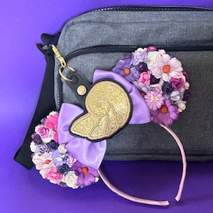 May include: A pair of decorative mouse ears with a seashell design and floral accents. The ears feature a gold glitter seashell, purple bows, and clusters of pink, purple, and white flowers. A black leather keychain with a gold clasp is attached.