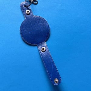 K&ouml;nnte beinhalten: Ein blauer Leder-Schl&uuml;sselanh&auml;nger mit rundem Design und langem, rechteckigem Riemen. Der Schl&uuml;sselanh&auml;nger hat silberfarbene Metallbeschl&auml;ge, einschlie&szlig;lich eines Verschlusses und Druckkn&ouml;pfen. Der Hintergrund ist einfarbig blau.