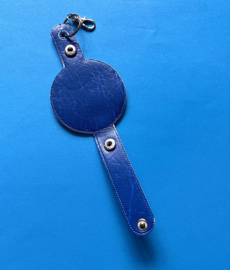 K&ouml;nnte beinhalten: Ein blauer Leder-Schl&uuml;sselanh&auml;nger mit rundem Design und langem, rechteckigem Riemen. Der Schl&uuml;sselanh&auml;nger hat silberfarbene Metallbeschl&auml;ge, einschlie&szlig;lich eines Verschlusses und Druckkn&ouml;pfen. Der Hintergrund ist einfarbig blau.