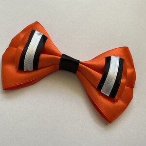 Peut inclure: Un nœud papillon orange vif avec des accents noirs et argentés. Le nœud papillon est fait d'un matériau semblable à du satin et comporte une bande centrale noire. Les extrémités du nœud papillon ont une rayure argentée bordée de rayures noires.