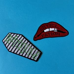 Può includere: Due toppe da stirare, una con una bara a strisce bianche e nere con il testo "Beetlejuice" in verde, e l'altra con una bocca rossa con denti bianchi e il testo "Anticipation" in bianco.