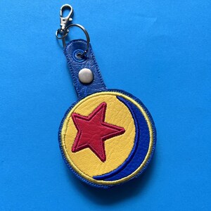 K&ouml;nnte beinhalten: Ein blauer Leder-Schl&uuml;sselanh&auml;nger mit einem silbernen Verschluss. Der Schl&uuml;sselanh&auml;nger hat ein kreisf&ouml;rmiges Emblem mit einem roten Stern in der Mitte, umgeben von einem gelben Hintergrund und einem blauen Halbmond-Design. Das Emblem ist mit gelbem Faden gen&auml;ht.