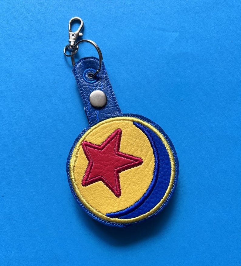 K&ouml;nnte beinhalten: Ein blauer Leder-Schl&uuml;sselanh&auml;nger mit einem silbernen Verschluss. Der Schl&uuml;sselanh&auml;nger hat ein kreisf&ouml;rmiges Emblem mit einem roten Stern in der Mitte, umgeben von einem gelben Hintergrund und einem blauen Halbmond-Design. Das Emblem ist mit gelbem Faden gen&auml;ht.