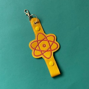 May include: Yellow keychain featuring a red atom symbol design. The keychain has a gold-coloured clasp and two yellow snap closures. The atom symbol is embroidered on the yellow leather. The background is teal.