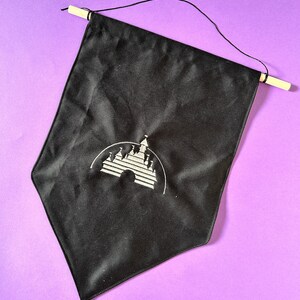 May include: A black fabric banner featuring a white embroidered image of the iconic Disney castle. The banner is suspended from a wooden dowel rod using a black string.