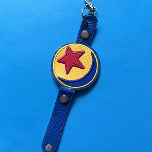 K&ouml;nnte beinhalten: Ein blauer Leder-Schl&uuml;sselanh&auml;nger mit einem gelben Kreis, einem roten Stern und einem blauen Halbmond-Design. Der Schl&uuml;sselanh&auml;nger hat einen silbernen Verschluss und zwei silberne Druckkn&ouml;pfe. Der Hintergrund ist einfarbig blau.