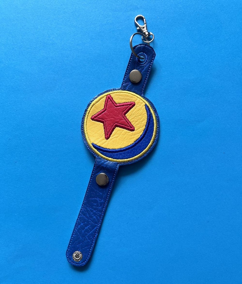 K&ouml;nnte beinhalten: Ein blauer Leder-Schl&uuml;sselanh&auml;nger mit einem gelben Kreis, einem roten Stern und einem blauen Halbmond-Design. Der Schl&uuml;sselanh&auml;nger hat einen silbernen Verschluss und zwei silberne Druckkn&ouml;pfe. Der Hintergrund ist einfarbig blau.