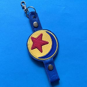 K&ouml;nnte beinhalten: Ein blauer Leder-Schl&uuml;sselanh&auml;nger mit einem kreisf&ouml;rmigen Emblem mit einem roten Stern auf gelbem Grund, umrahmt von einem blauen Halbmond. Der Schl&uuml;sselanh&auml;nger hat einen silbernen Verschluss und einen Druckknopfverschluss.