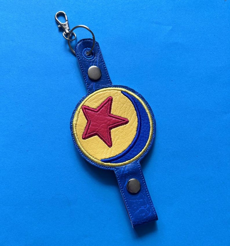 K&ouml;nnte beinhalten: Ein blauer Leder-Schl&uuml;sselanh&auml;nger mit einem kreisf&ouml;rmigen Emblem mit einem roten Stern auf gelbem Grund, umrahmt von einem blauen Halbmond. Der Schl&uuml;sselanh&auml;nger hat einen silbernen Verschluss und einen Druckknopfverschluss.