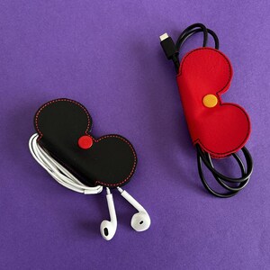 May include: Two cable organisers, one red and one black, with a stitched design. The red organiser is shaped like a heart and the black organiser is shaped like a mouse head. Both organisers are made of felt and have a small button on the front.