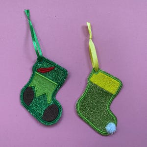 Puede incluir: Dos adornos de calcetín navideño. Uno es verde con un acento rojo y talón y puntera marrones. El otro es verde con un puño amarillo y un pompón blanco. Ambos tienen una cinta para colgar.