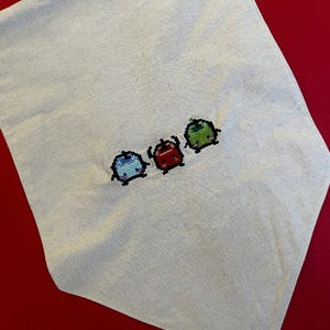 Puede incluir: Un artículo de tela de color claro con un diseño de punto de cruz de tres pequeñas criaturas coloridas. Las criaturas son azules, rojas y verdes, cada una con contornos negros. El fondo es de color neutro, sobre un fondo rojo.