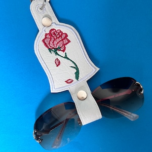 K&ouml;nnte beinhalten: Ein wei&szlig;er Sonnenbrillenhalter aus Kunstleder mit einem roten Rosendesign, einem gr&uuml;nen Stiel und einem silbernen Schl&uuml;sselring. Der Halter ist an einer Sonnenbrille mit dunklen Gl&auml;sern und silbernen Rahmen befestigt. Der Hintergrund ist einfarbig blau.