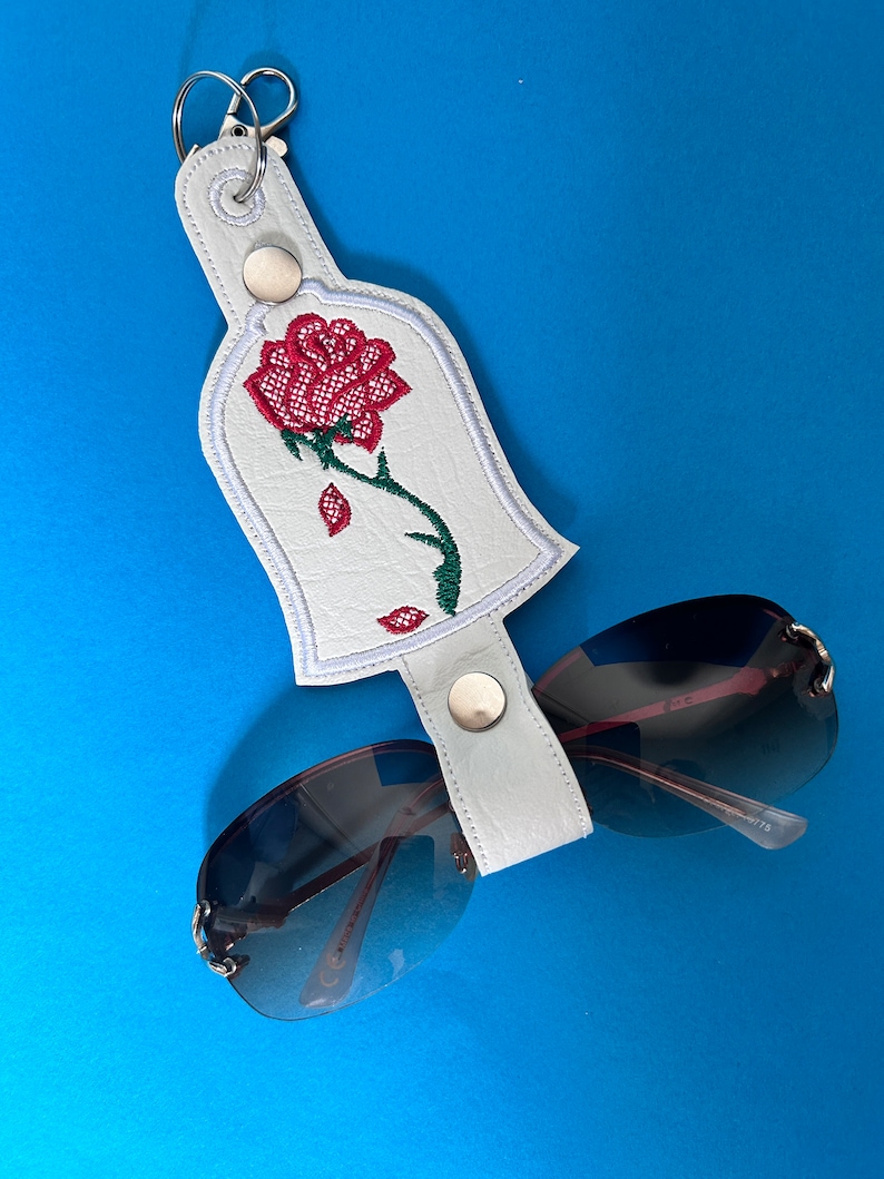 K&ouml;nnte beinhalten: Ein wei&szlig;er Sonnenbrillenhalter aus Kunstleder mit einem roten Rosendesign, einem gr&uuml;nen Stiel und einem silbernen Schl&uuml;sselring. Der Halter ist an einer Sonnenbrille mit dunklen Gl&auml;sern und silbernen Rahmen befestigt. Der Hintergrund ist einfarbig blau.