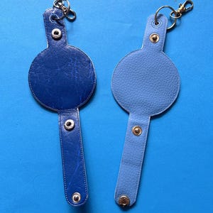 K&ouml;nnte beinhalten: Zwei blaue Leder-Schl&uuml;sselanh&auml;nger. Einer ist dunkelblau mit strukturierter Oberfl&auml;che, der andere hellblau mit glatter Textur. Jeder Schl&uuml;sselanh&auml;nger hat eine kreisf&ouml;rmige Oberseite und einen rechteckigen Riemen mit Druckkn&ouml;pfen und einem Metallring.