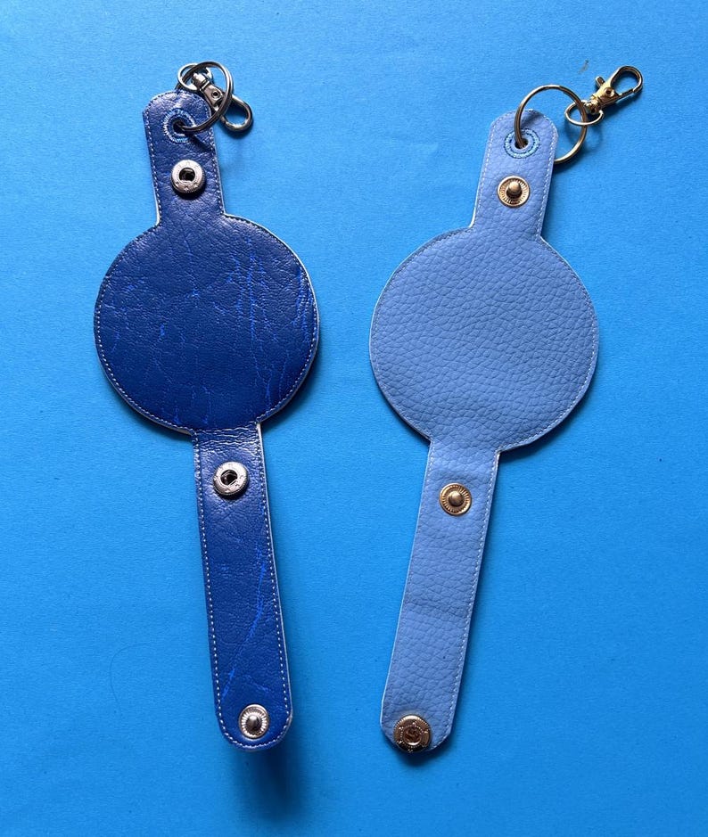 K&ouml;nnte beinhalten: Zwei blaue Leder-Schl&uuml;sselanh&auml;nger. Einer ist dunkelblau mit strukturierter Oberfl&auml;che, der andere hellblau mit glatter Textur. Jeder Schl&uuml;sselanh&auml;nger hat eine kreisf&ouml;rmige Oberseite und einen rechteckigen Riemen mit Druckkn&ouml;pfen und einem Metallring.