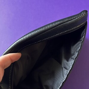 May include: A black leather zippered pouch with a black fabric lining. The pouch is open and the lining is visible.