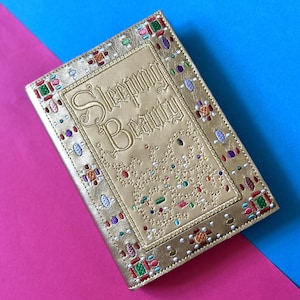 Peut inclure: Une couverture de livre dorée avec le titre "Sleeping Beauty" brodé dans une police décorative. La couverture est ornée de détails brodés colorés et d'un motif pointillé.