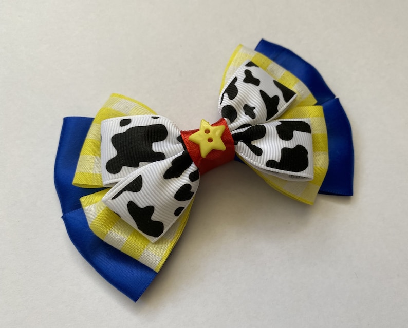 Pu&ograve; includere: Un fiocco giallo, bianco e blu con un nastro a stampa di mucca bianco e nero e un bottone rosso con una stella gialla.