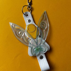 May include: A white and yellow fairy keyring with clear, iridescent wings and a silver clasp. The fairy has a flower-like design on its chest and is attached to a white strap. The background is a solid yellow.