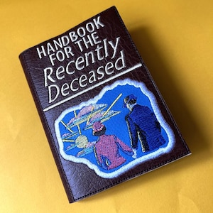 May include: A brown leather-bound book with the title "Handbook for the Recently Deceased" embroidered in white thread. The cover features an embroidered patch of two figures walking towards a bright light in the sky.